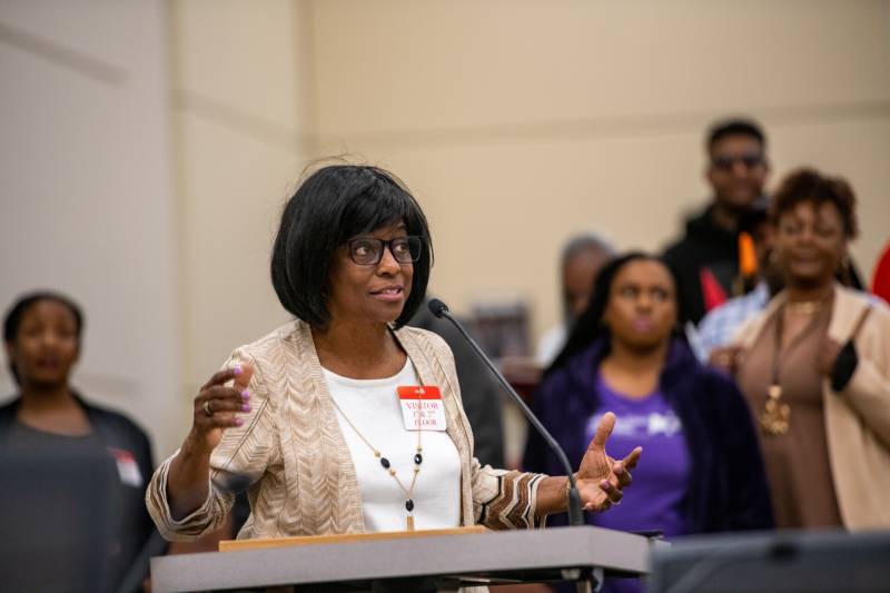 A middle-aged African American woman speaks at a dais with people behind her.