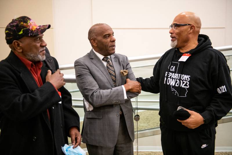 Three older African American men stand having a conversation. The man on the far left, with a neat gray beard, wears a black baseball cap with colorful splotches of pink, yellow and green, watching the other two men speak. The man in the middle, mostly bald, wears a gray suit of a very light plaid, an ochre pocket handkerchief, and an ochre-and-navy-striped tie. He faces the last man, on the far right, who is speaking and seems to be resting his right hand on the back of the man in the suit. He is the tallest, bald and with a white goatee and glasses, and a black hoodie with an outline of California in white that says "CA Reparations Now 2023," and a sticker name tag on his chest.