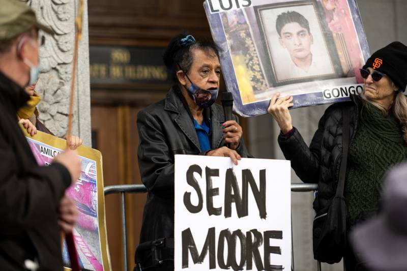 An older Black woman, wearing a blue and purple cloth mask pulled to her chin, and wearing a long black trench coat and a bright blue blouse, speaks into a microphone, holding a sign that says "Sean Moore." The wooden doors of the building and gold text on a transom window above them are blurred in the background.