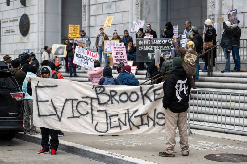 People gathered outside a court building holding banners, with a large banner in foreground that reads "Evict Brooke Jenkins"