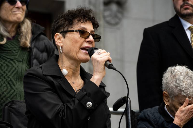 A woman speaks into a microphone with people standing around her and a court building behind her.
