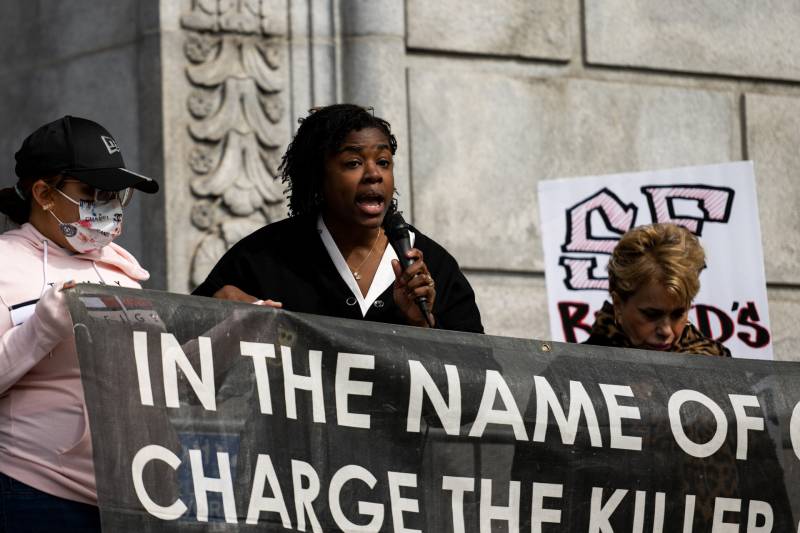 A younger African American woman, with shoulder-length black hair and a white blouse beneath a black cardigan, speaks into a microphone she holds, standing behind a large black banner that reads in white text, "In the name of [out of frame] charge the killer."