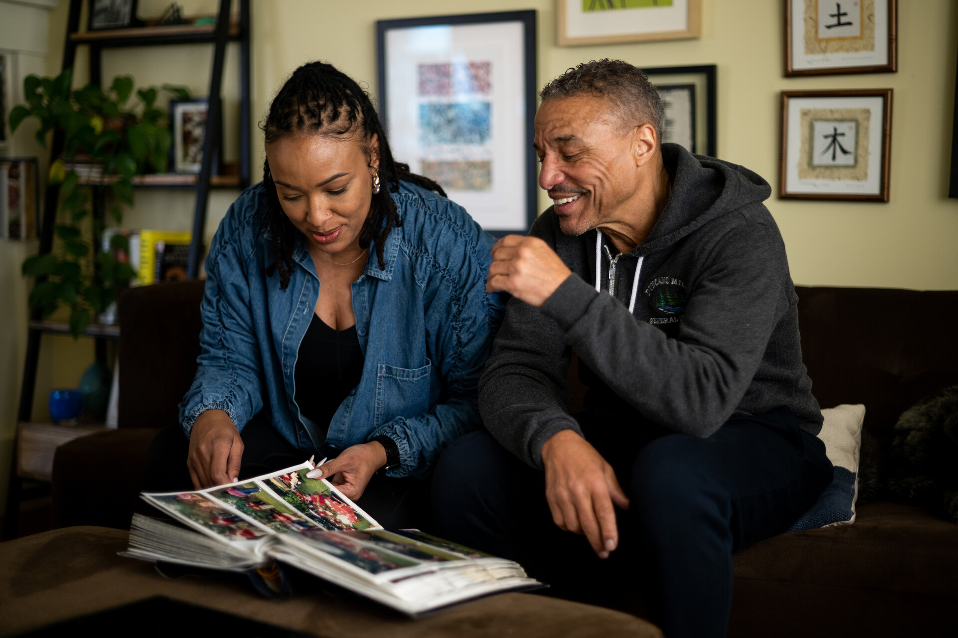 A Black woman wearing a jean, long-sleeved shirt and black T-shirt underneath sits on a couch inside a living room with her father to the right. He wears a gray, hooded sweatshirt. The two smile as they look down at a photo album together.