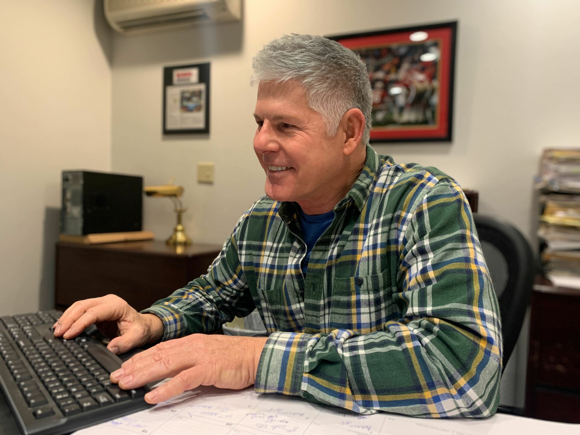 A middle-aged white man in a green plaid shirt sits smiling at a desk typing on a computer keyboard.