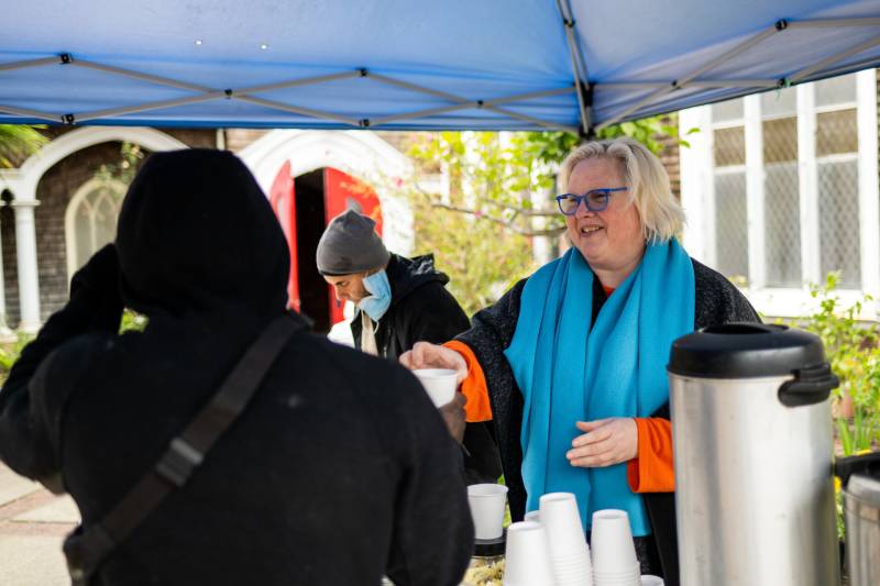 An older white woman with a blue shawl smiles as she hands a white cup to a person with a hoodie seen from behind, under a tent with another person in the background.