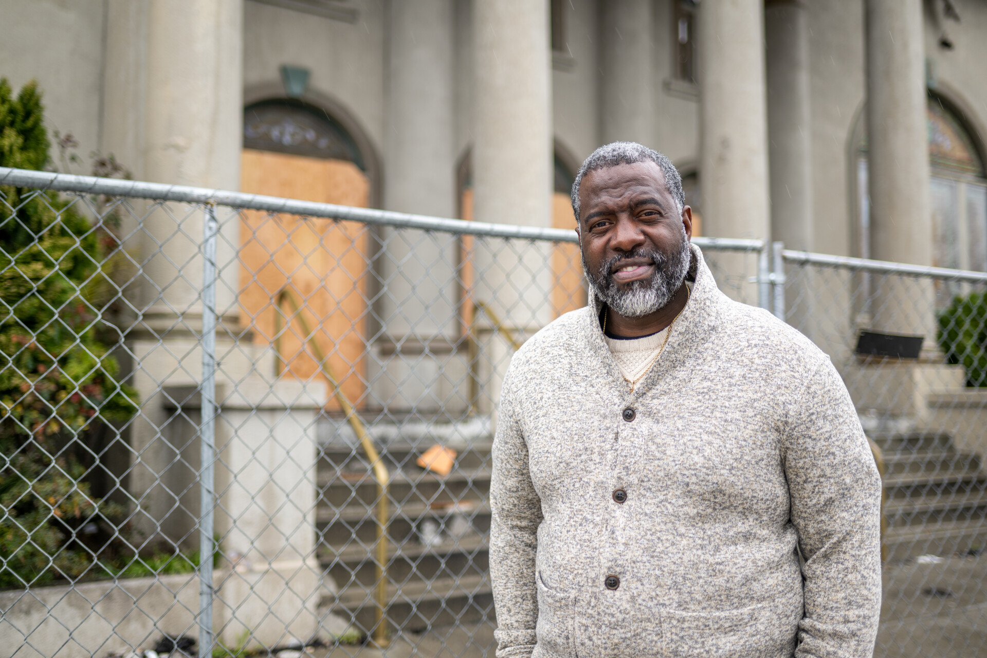 A Black man with salt-and-pepper hair and a full beard wearing a light grey cardigan stands outside a building with a fence behind him.