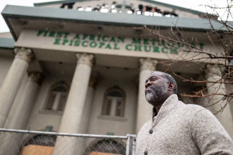 An African American man wearing a light grey sweater vest stands outside a building with a sign that reads "First African Methodist Episcopal Church."