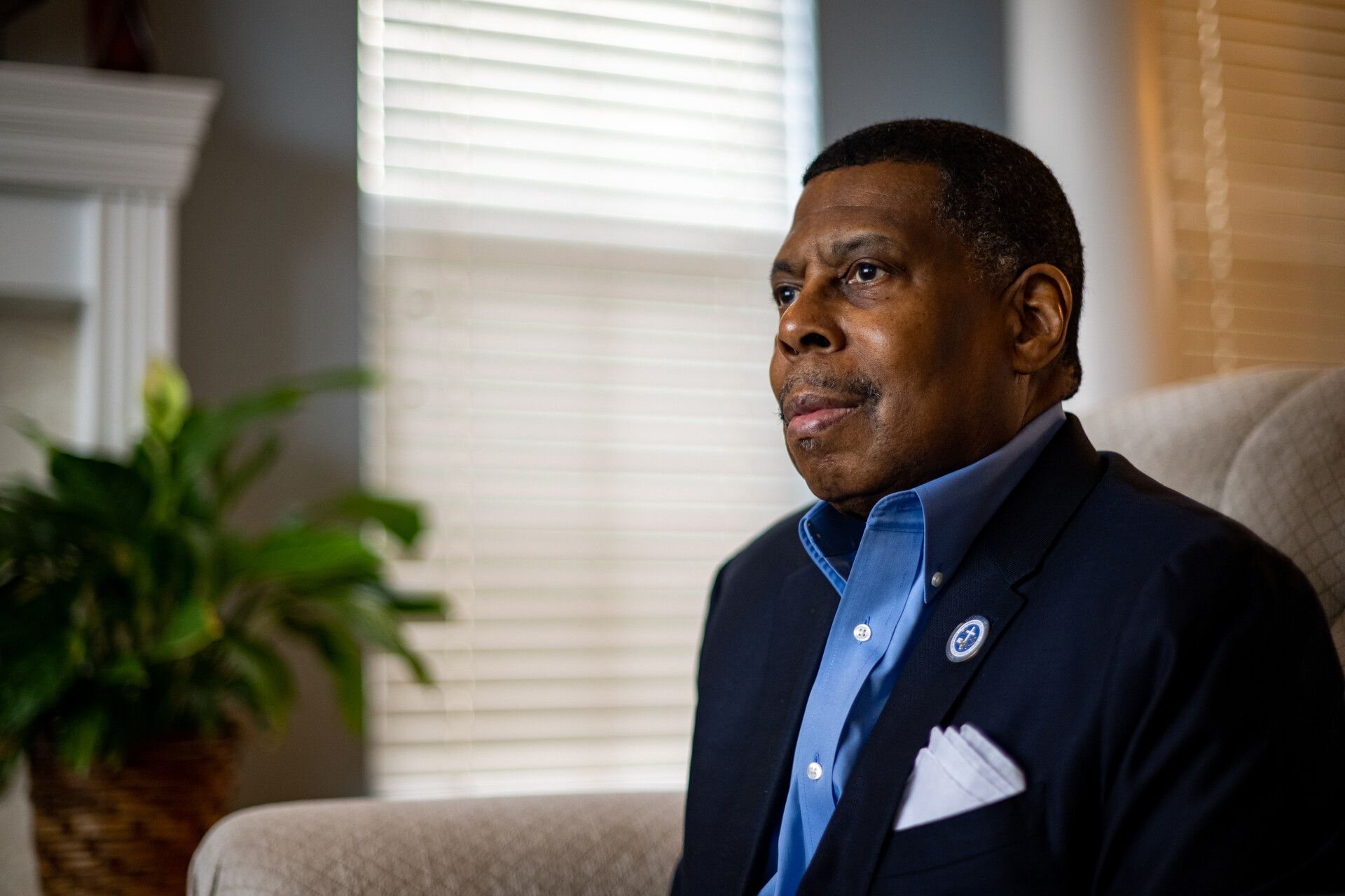 A distinguished middle-aged Black man wearing a blue collared shirt and blue jacket sits in a well-lit living room, his eyes looking into the distance