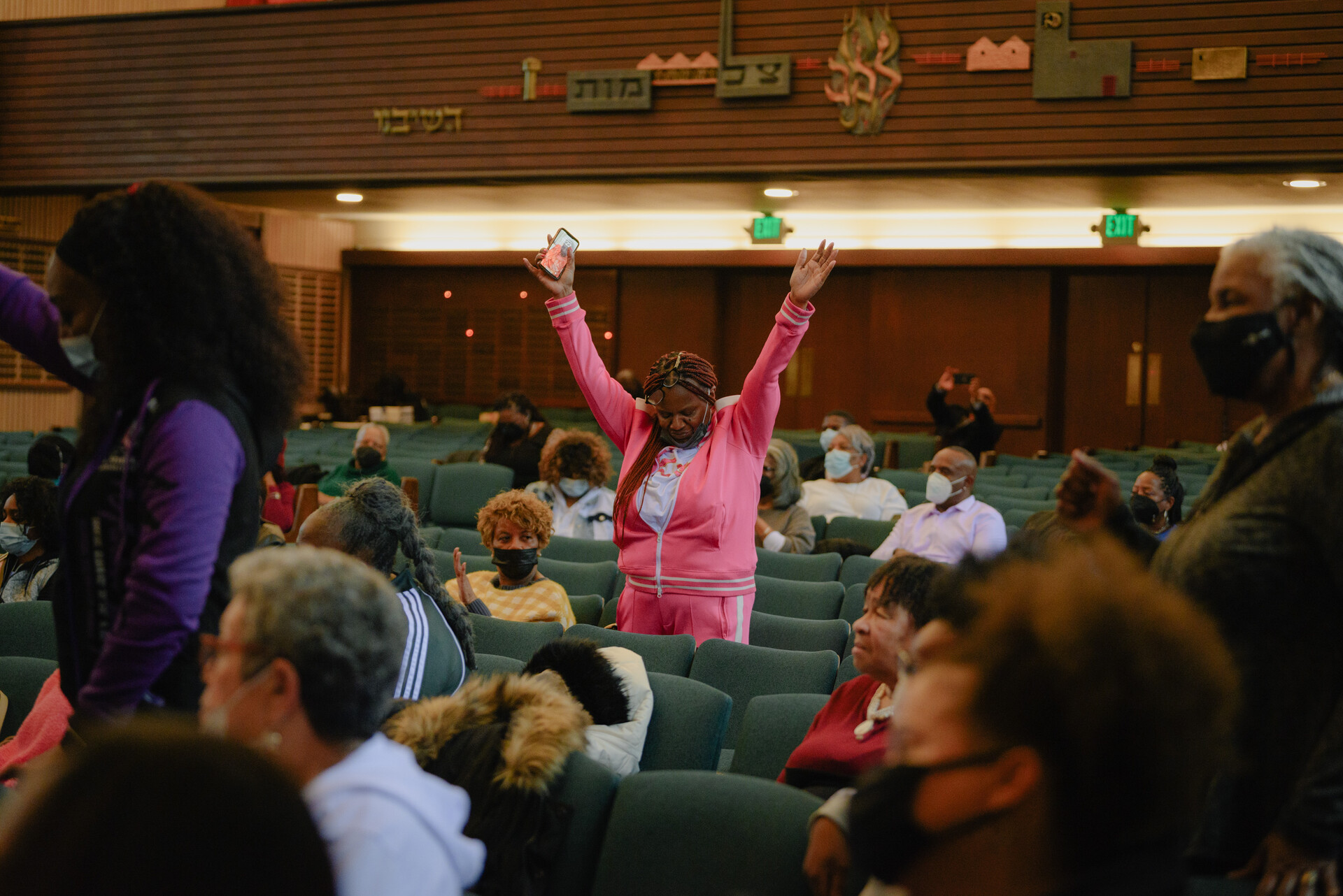 An African American woman wearing a pink jumpsuit stands with both hands raised among several people who are seated in a building.