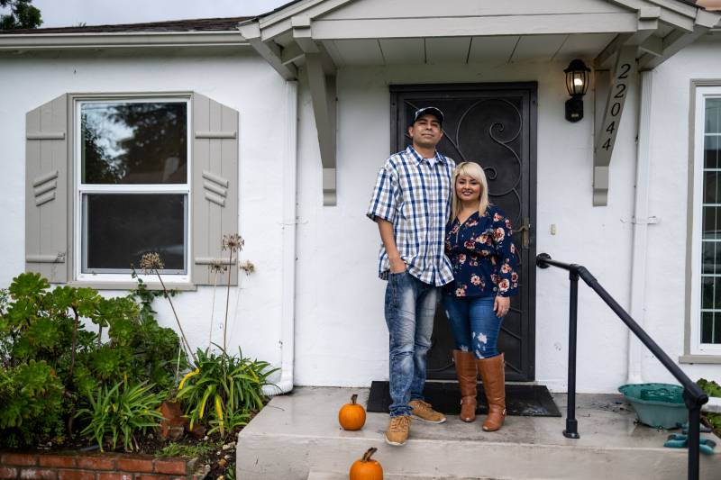 A tall man stands with his arm around a shorter woman, in front of the front door a small white house, with small pumpkins lining the front steps.