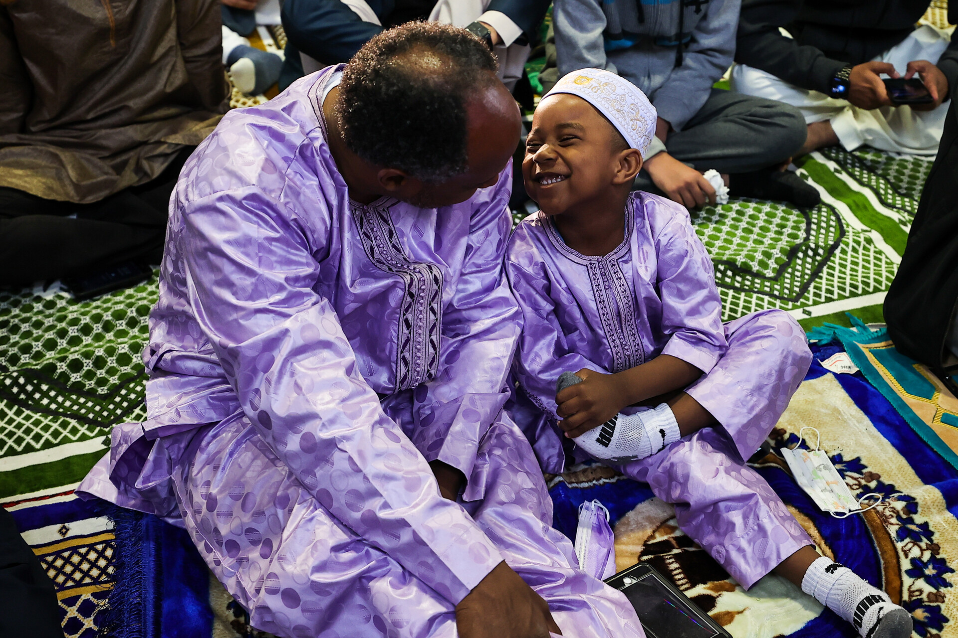 A man and a young boy, both with darker skin, wear traditional long sleeved purple shirts and pants, sitting down on a prayer mat. The boy is smiling broadly as the man looks down at him.