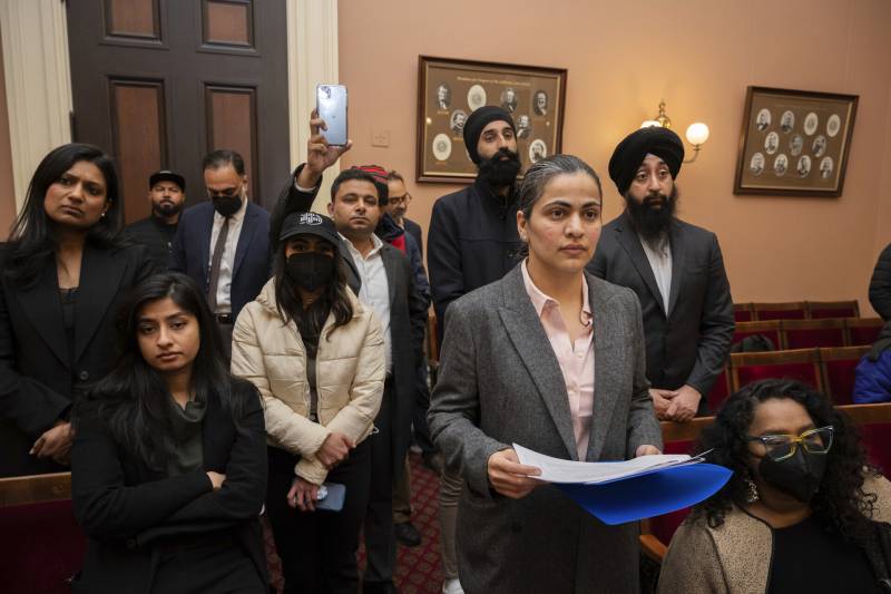 Men and women in business attire stand in a courtroom in Sacramento, California. One man holds up his cell phone One woman in a gray suit with pink button-up shirt, holds a blue folder with papers inside as she looks past the camera.