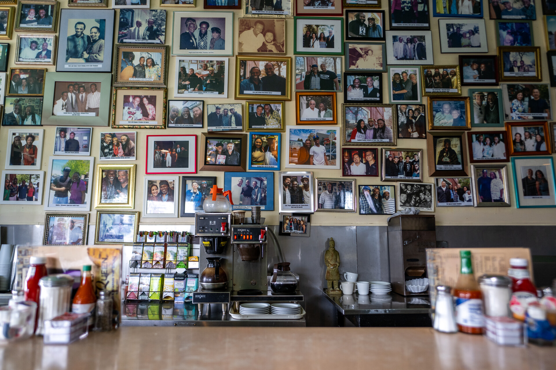 A close up of a diner counter with hot sauce bottles, glass sugar containers, salt and pepper shakers, packets of jelly and plastic bottles of ketchup neatly arranged. In the background, a large collage of individually framed photos decorate the wall leaving no room between each frame. In the center, coffee pots warm on the coffee station.