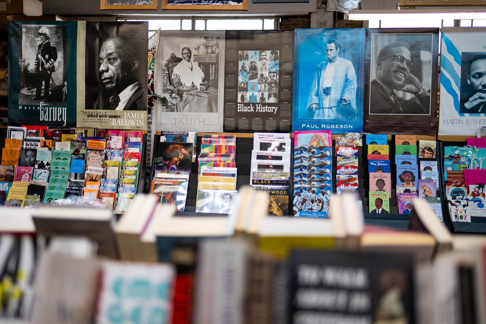 Inside a bookstore, a wall is covered in colorful imagery and black and white posters of historic figures such as James Baldwin, Paul Robeson, Malcolm X and Martin Luther King Jr.