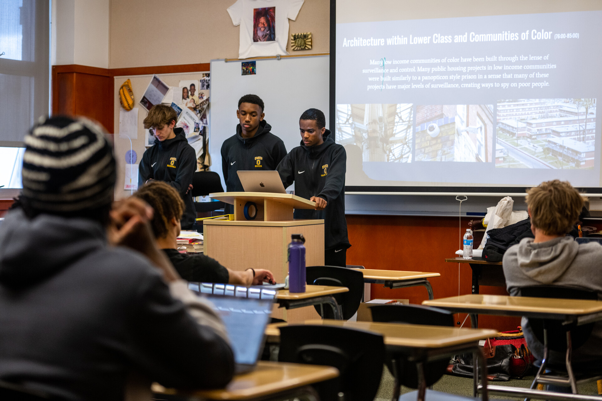 Students sit at desks inside a classroom. A projector displays a presentation. Three high school boys stand at the podium in front of the classroom ready to speak.