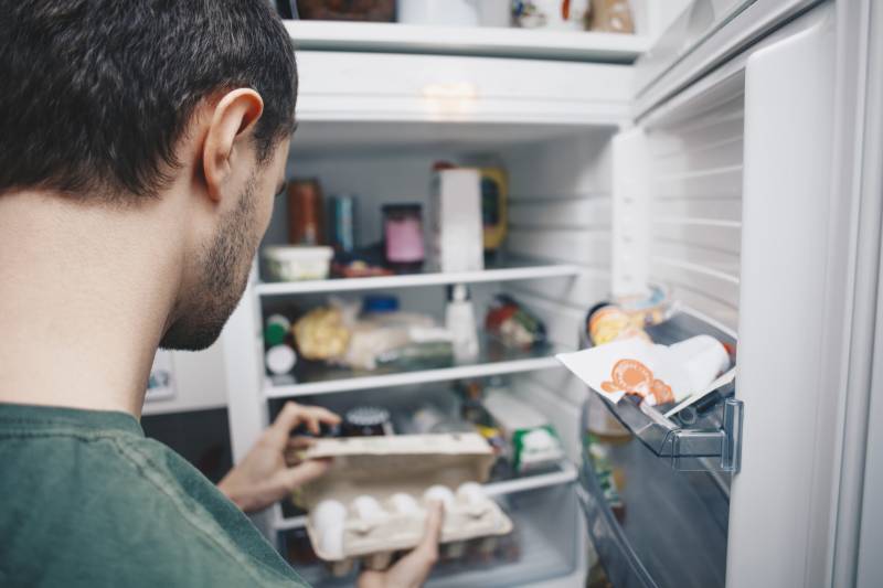 A person photographed from behind their head, looking in a fridge and holding some eggs in their hands.