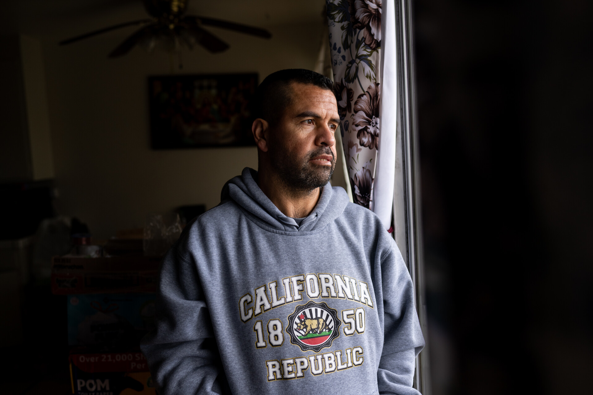 A man with short, dark hair and a five o'clock shadow wears a gray hooded sweatshirt that reads "California 1850 Republic" as he stares out the window from his apartment. The sunlight illuminates his serious face.
