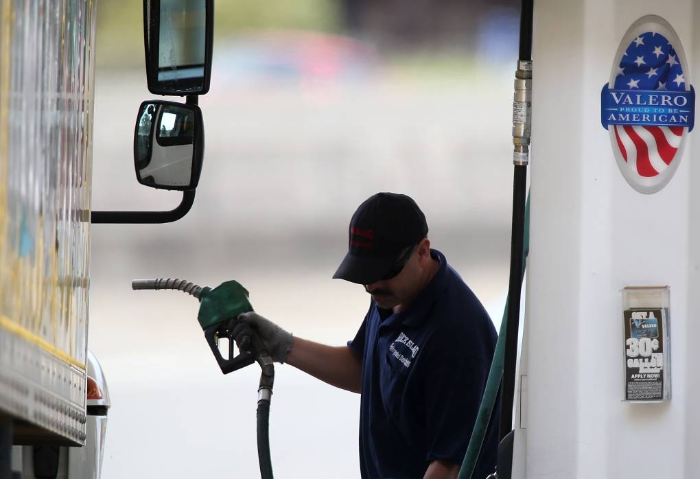 A man at a gas station prepares to fill up his diesel vehicle. He wears a ballcap and blue collared work shirt.