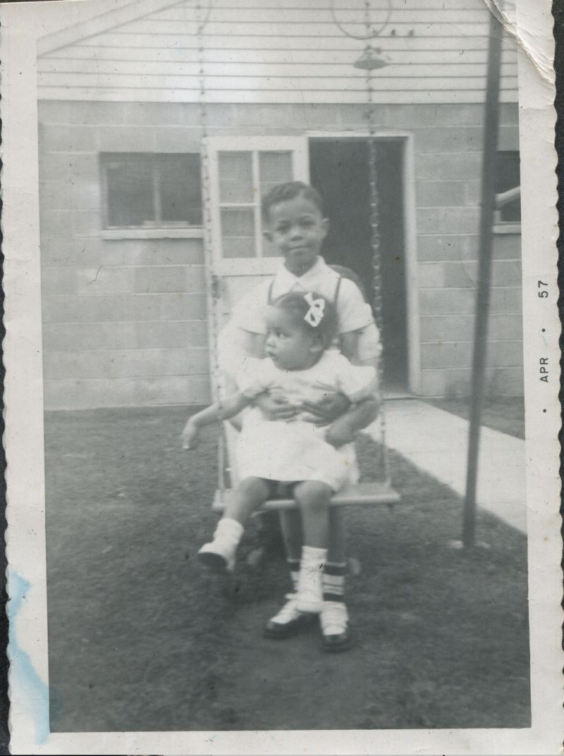 A vintage black and white of a young boy holding a baby girl while sitting on a swing set outdoors.