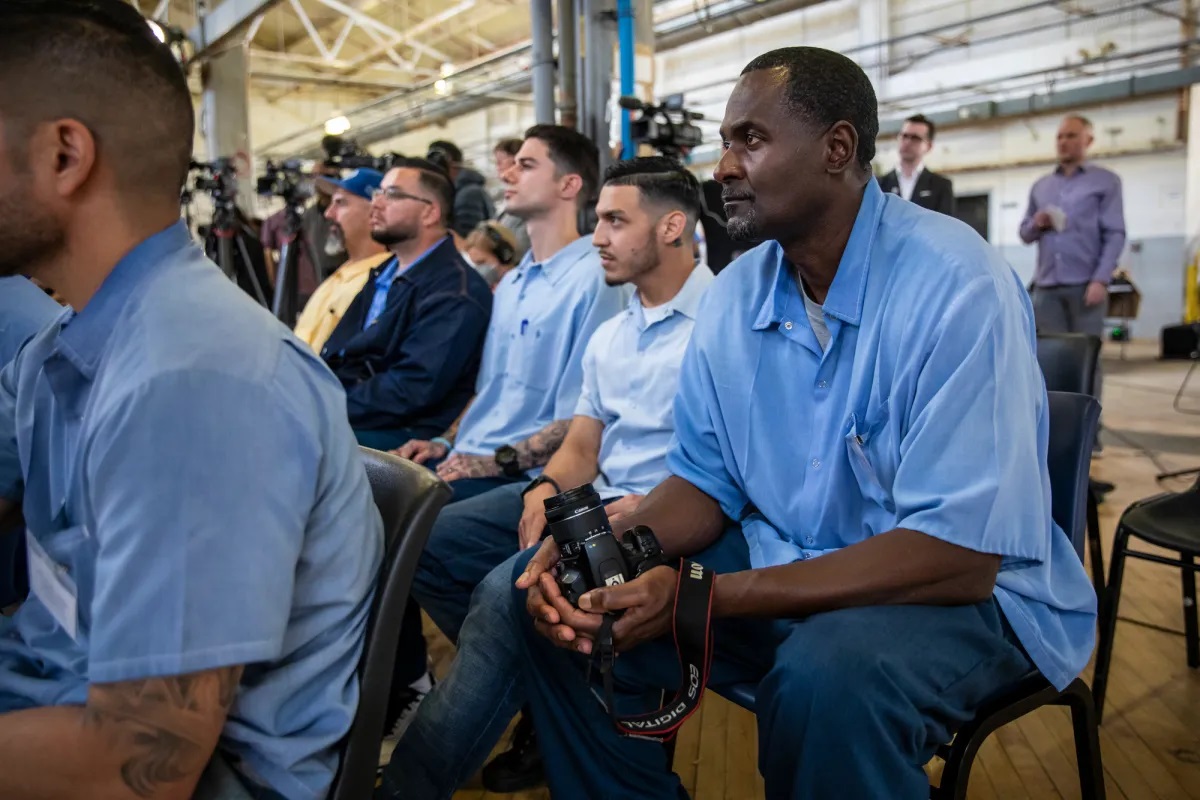 Prisoners of San Quentin, wearing light blue shirts and navy pants, sit on chairs in rows observing a speech by Gov. Gavin Newsom. One man in the foreground holds a digital camera as he looks onward.