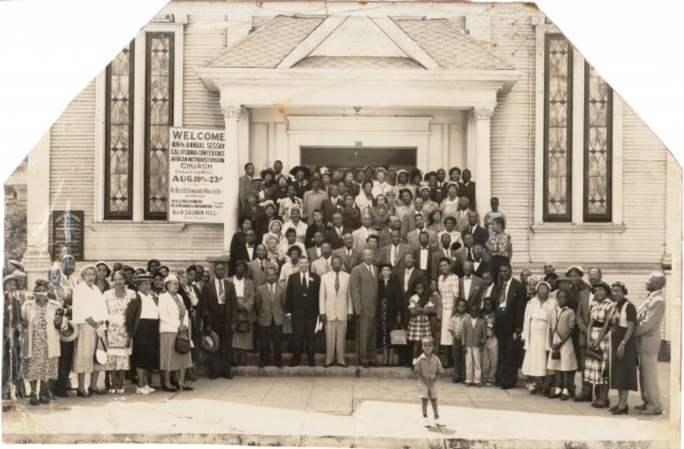 An archival photo of a large group of African American men, women, and children standing in front of a building.