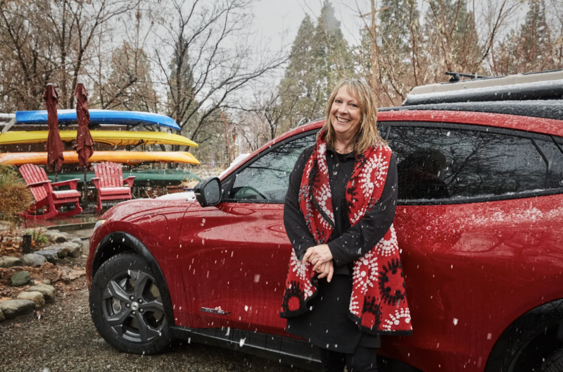 A white woman wearing a red and black jacket standing next to a red vehicle outside as it snows with water vessels stacked in the background.