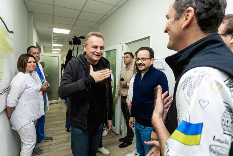 grinning white man wearing dark clothing extends his hand towards another smiling white man closer to the camera who is wearing a jacket with the Ukrainian flag on it