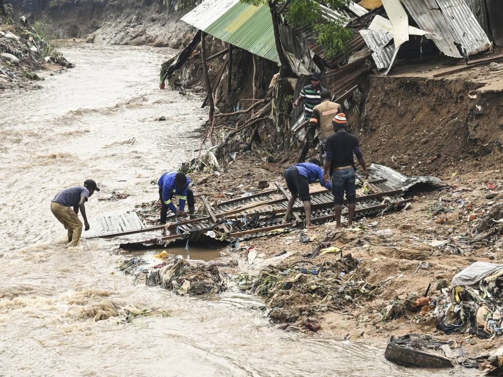 A massive stream of brown water flows rapidly between an embankment as six men work to salvage wood and metal scraps from a recent flood in Africa.