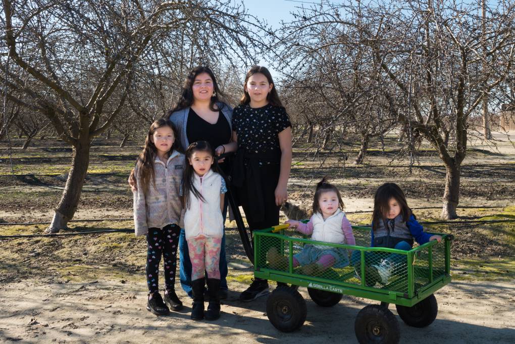 A Latina family with mother and four daughters and one son of various ages, from toddler to teenager, standing in an almond orchard. The two youngest children are seated in a green-painted metal wagon.