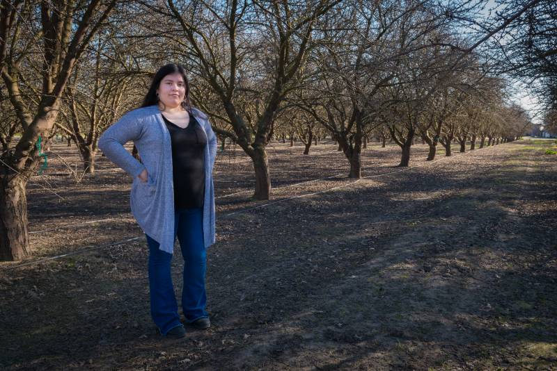 A Latina woman with a blue cardigan and a black shirt looks at the camera, arms akimbo, standing in a leafless orchard.