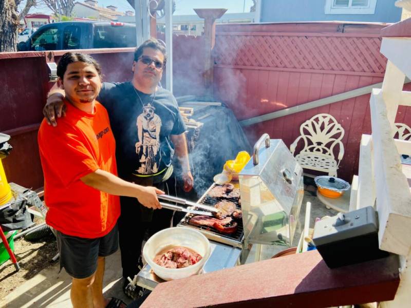 A man wearing a black t-shirt and sun glasses has his arm around a younger man wearing an orange t-shirt who is holding a cooking tool over a grill outside.