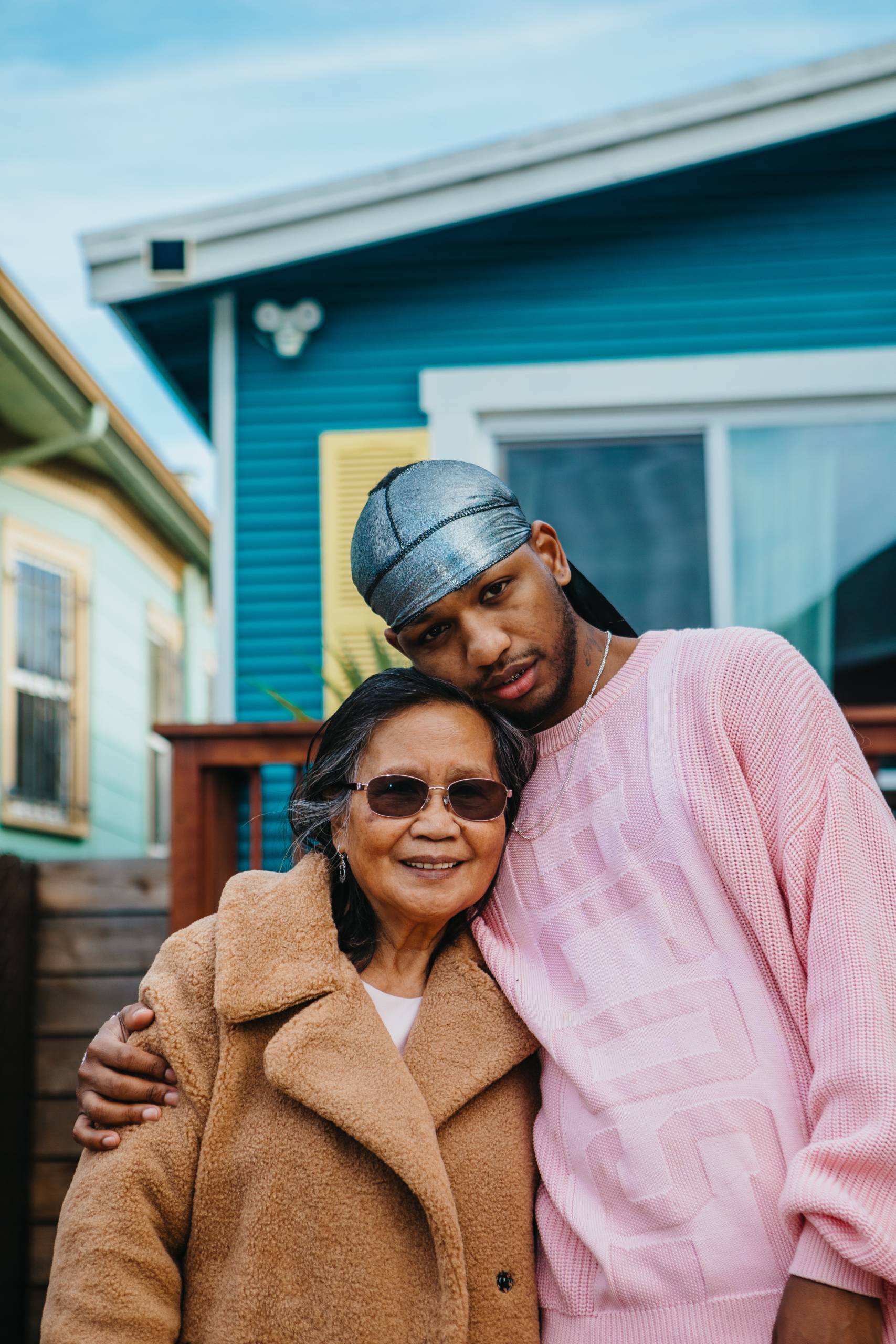 A Filipina grandmother in a fuzzy, brown coat and wearing sunglasses smiles as she poses for a photo with her grandson, who towers above her and lays his cheek on top of his head, wears a pink, knitted sweater and silver durag.
