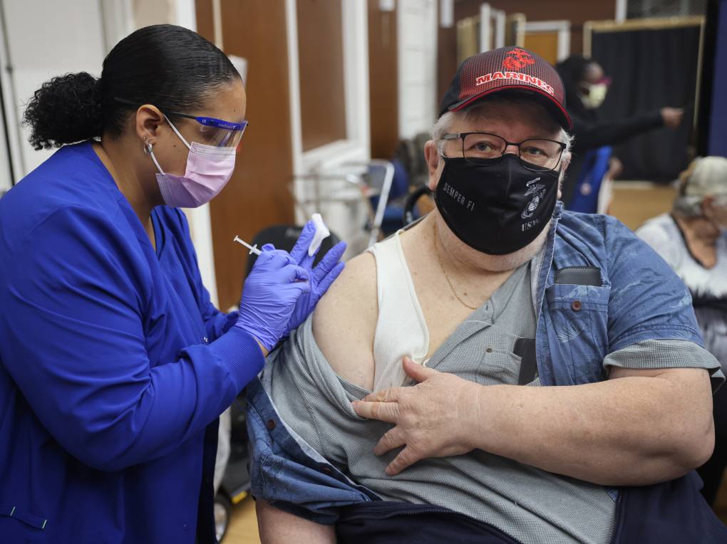 A nurse in blue scrubs and purple latex gloves wears a mask and goggles as she administers a needle into the arm of a patient who moves his denim shirt off of his shoulder for her. He wears a black mask and his gray hair is seen from under his black baseball cap. More patients and nurses are seen in the background from inside a clinic setting.