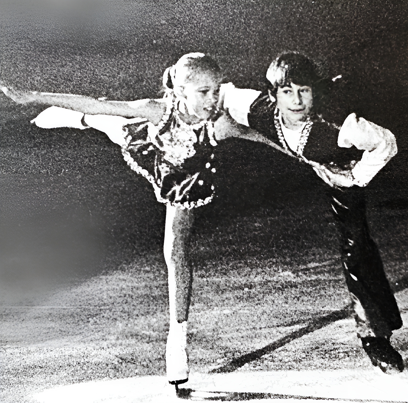 Two child figure skaters, one girl with a blond ponytail, and one boy with dark, short hair, glide on the ice together in a black and white image.