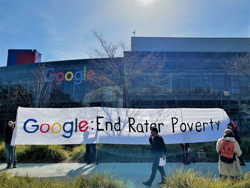 A view of Google headquarters with a banner in front of it that reads "Google: End Rater Poverty" and a person standing in front of the banner.