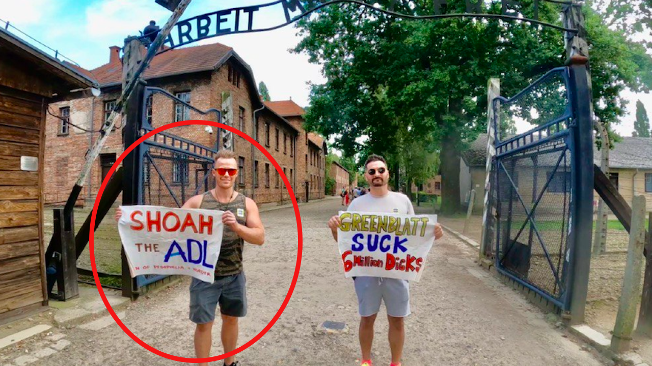 Two white men wearing sun glasses, a sleeveless t shirt and shorts hold signs while standing outside in the street with buildings to the left and trees to the right. The Signs read "Shoah the ADL" and "Greenblatt suck 6 million dicks." A red circle is around the man on the left.