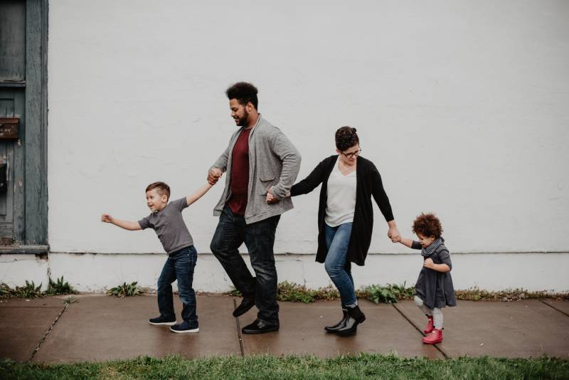 A family of four -- two adult parents or caregivers, and two children -- are photographed skipping along a wet street, holding hands.