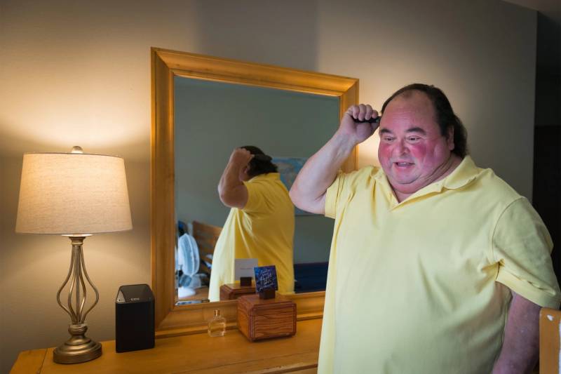 A white middle aged man combs his hair in front of a mirror in a bedroom with a lamp, a speaker, a bottle of eau de toilette anfd a box with a card on a dresser in front of the mirror.
