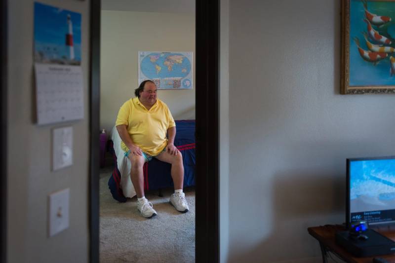 A middle aged white man wearing a yellow polo shirt and shorts with white sneakers sits on the edge of his bed, seen through the doorway of another room where there is a computer to the side.