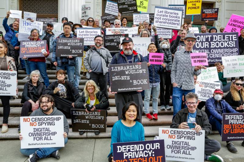 A group of about 40 people stand on steps in front of a building holding numerous signs, a prominent one reading 'librarians wouldn't delete a book'