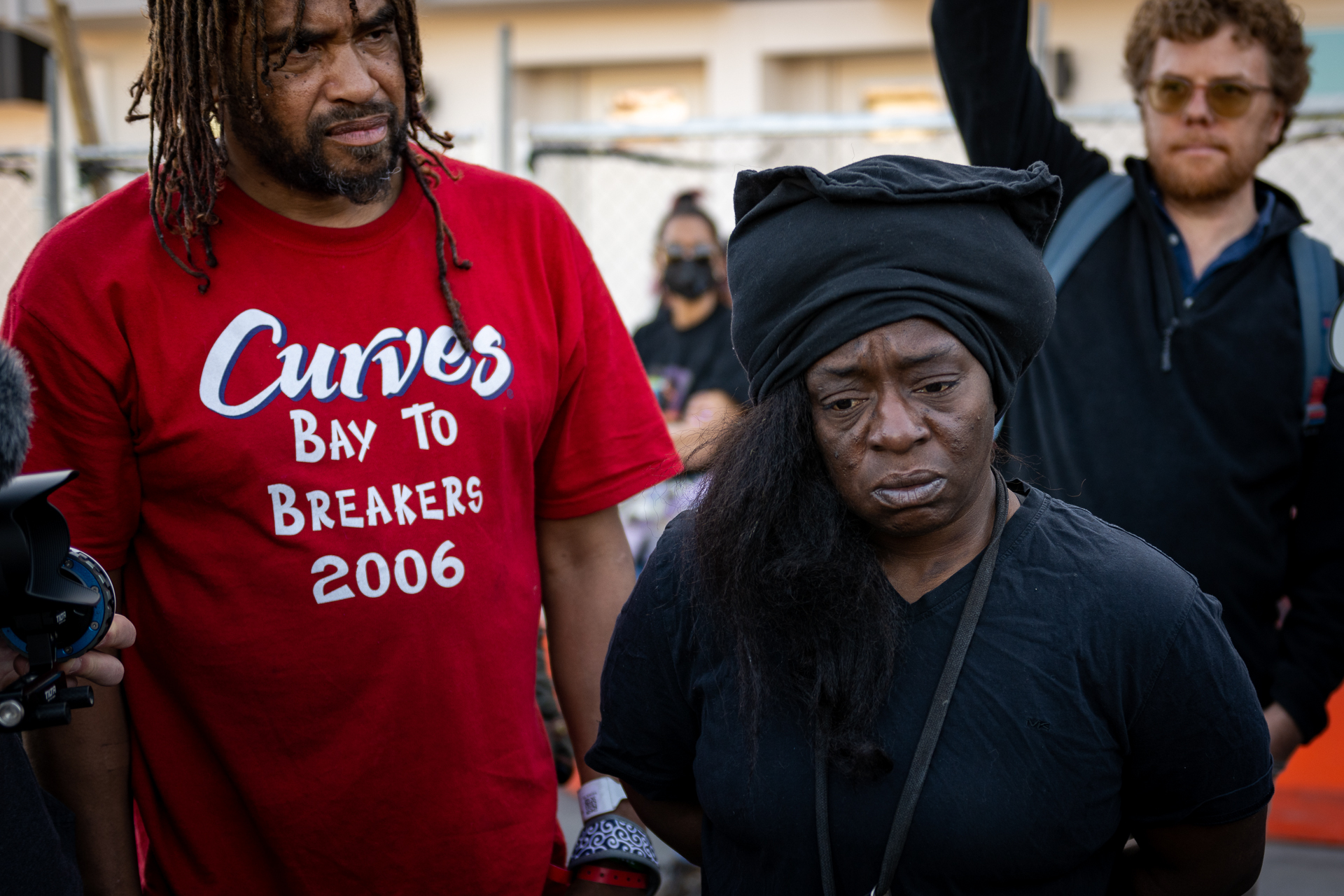 A Black woman with a black headdress and T-shirt looks heartbroken as she stares downward. Two men, one Black and one white-presenting, are pictured behind her.