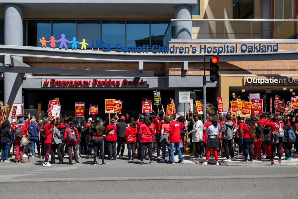 Hundreds of protestors in red T-shirts hold red and yellow picket signs outside of UCSF Benioff Children's Hospital Oakland.