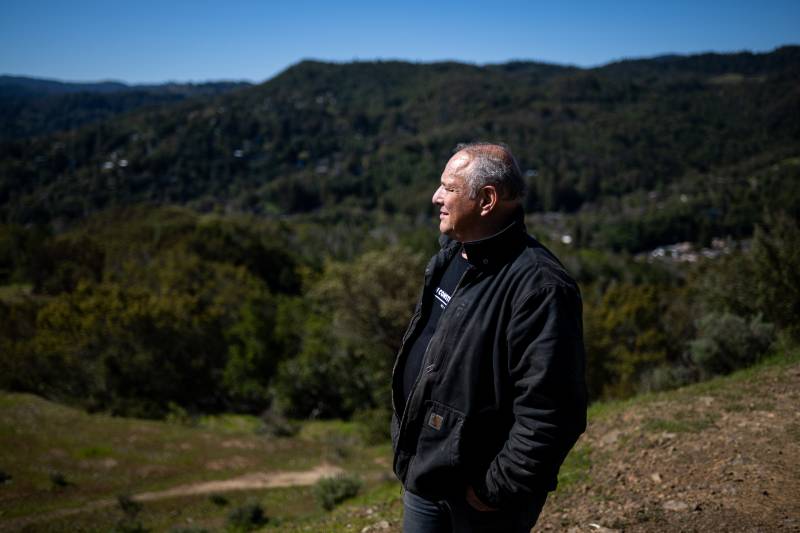 A man with gray hair and a black jacket and T-shirt looks off into the distance on land that's covered in green trees with blue skies in the background.