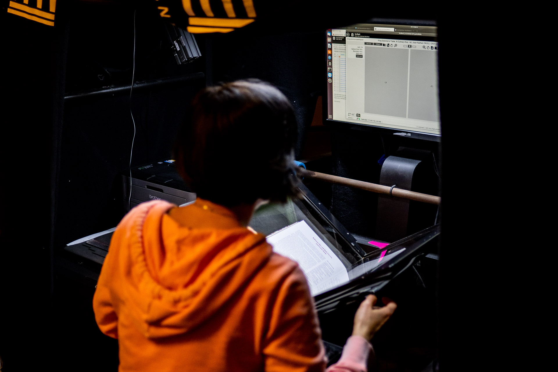 Someone wearing a bright orange hoodie sits at an archiving station holding an open book and facing a computer screen