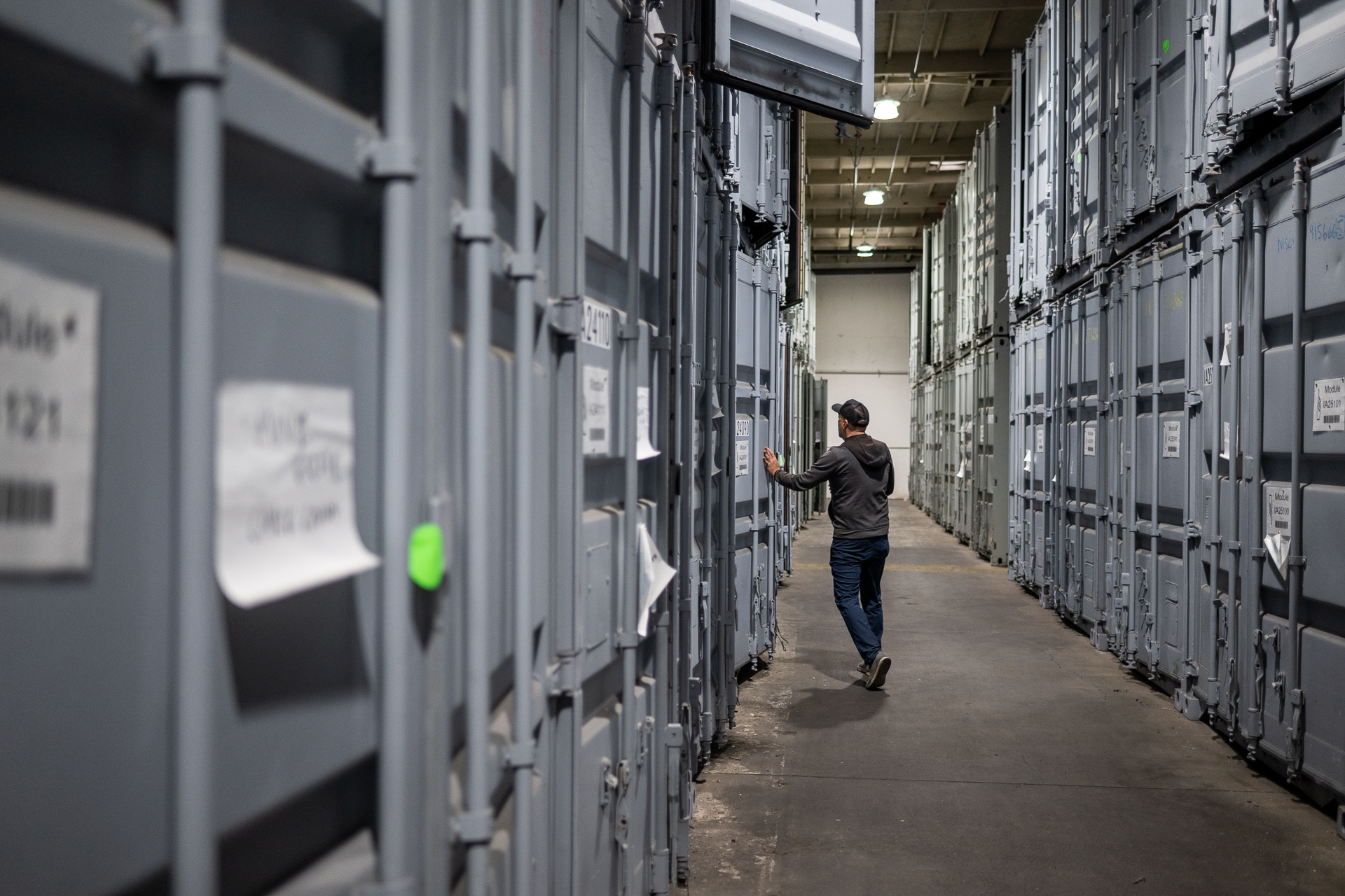 A man in the distance stands in a walkway between two huge walls of grey storage containers stacked on top of each other inside what appears to be a massive warehouse