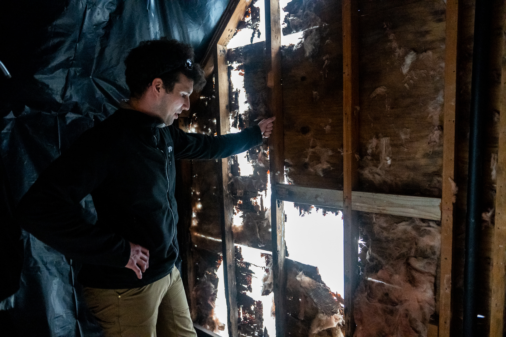 A man in a black, zip-up sweater stands inside a dilapidated house where the walls have enormous holes and is in need of heavy repairs. He peers down through one of the openings with one hand on his right hip.