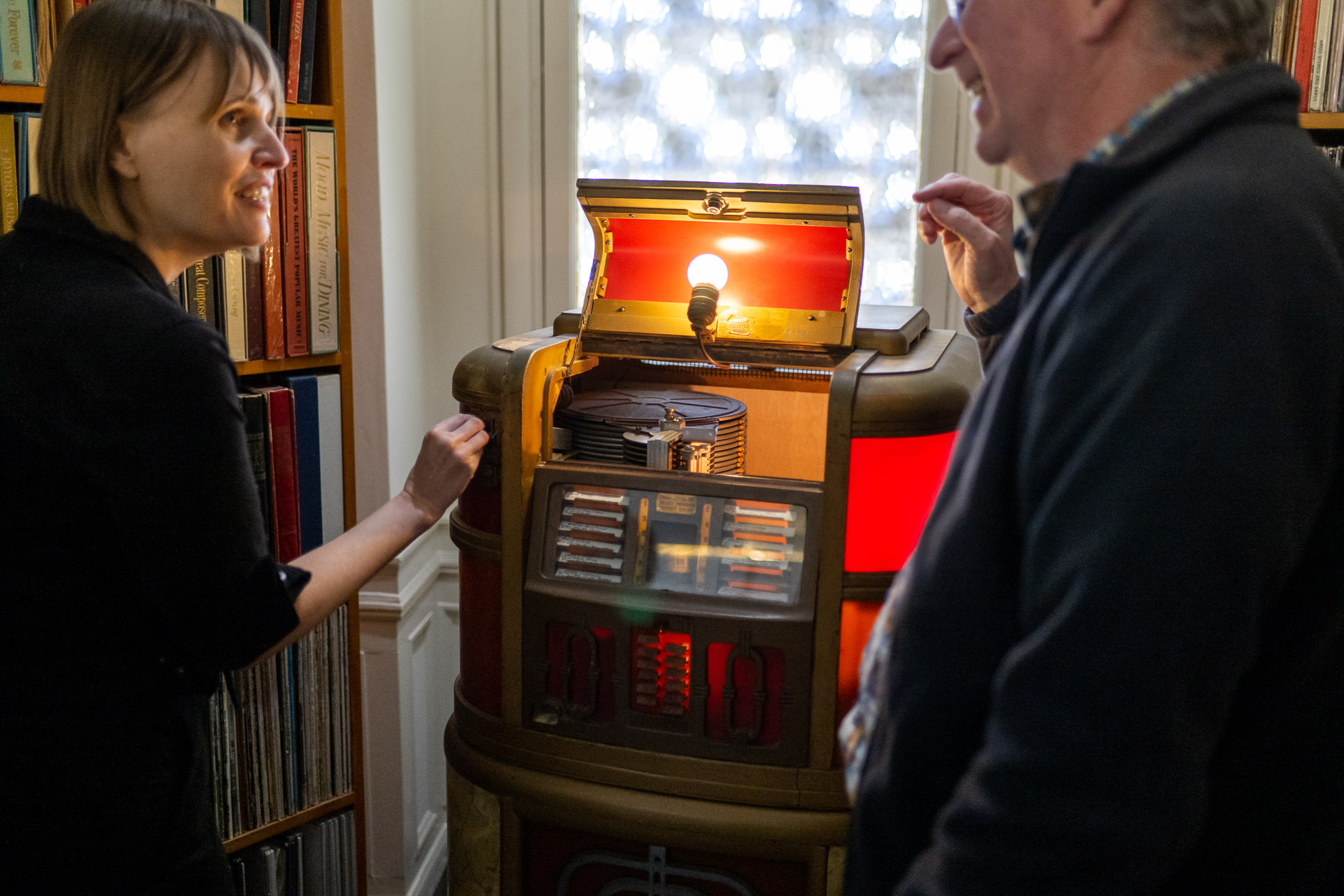 A white woman with short brown hair stands facing a middle-aged white man, both smiling and engaged in conversation, with an old time record player in the background within a corridor which appears to be lined with vinyl records
