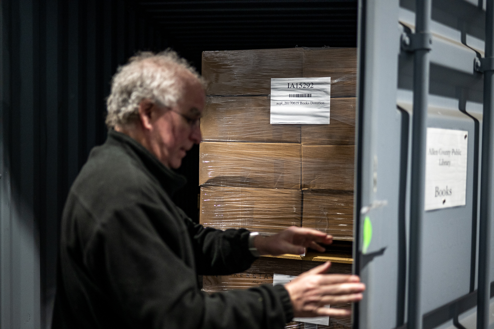 An older white man with grey-white hair wearing a dark sweater reaches out to close a grey metallic door as huge cardboard boxes labeled as containing books sit in the background