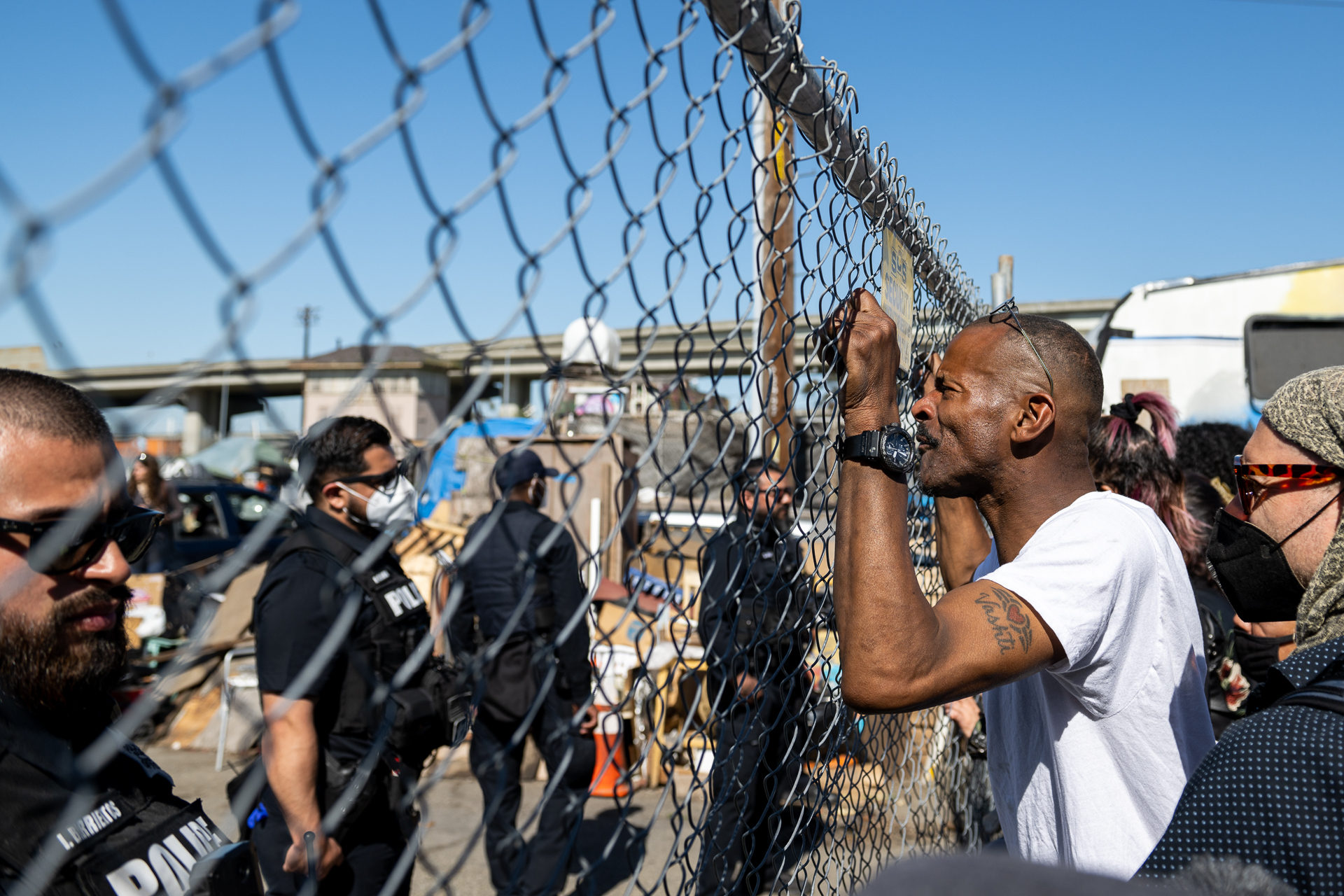 A Black man looks through a chain link fence at police officers on the other side of it. The man has a crowd of people behind him as his fingers rest in between the fence coils.