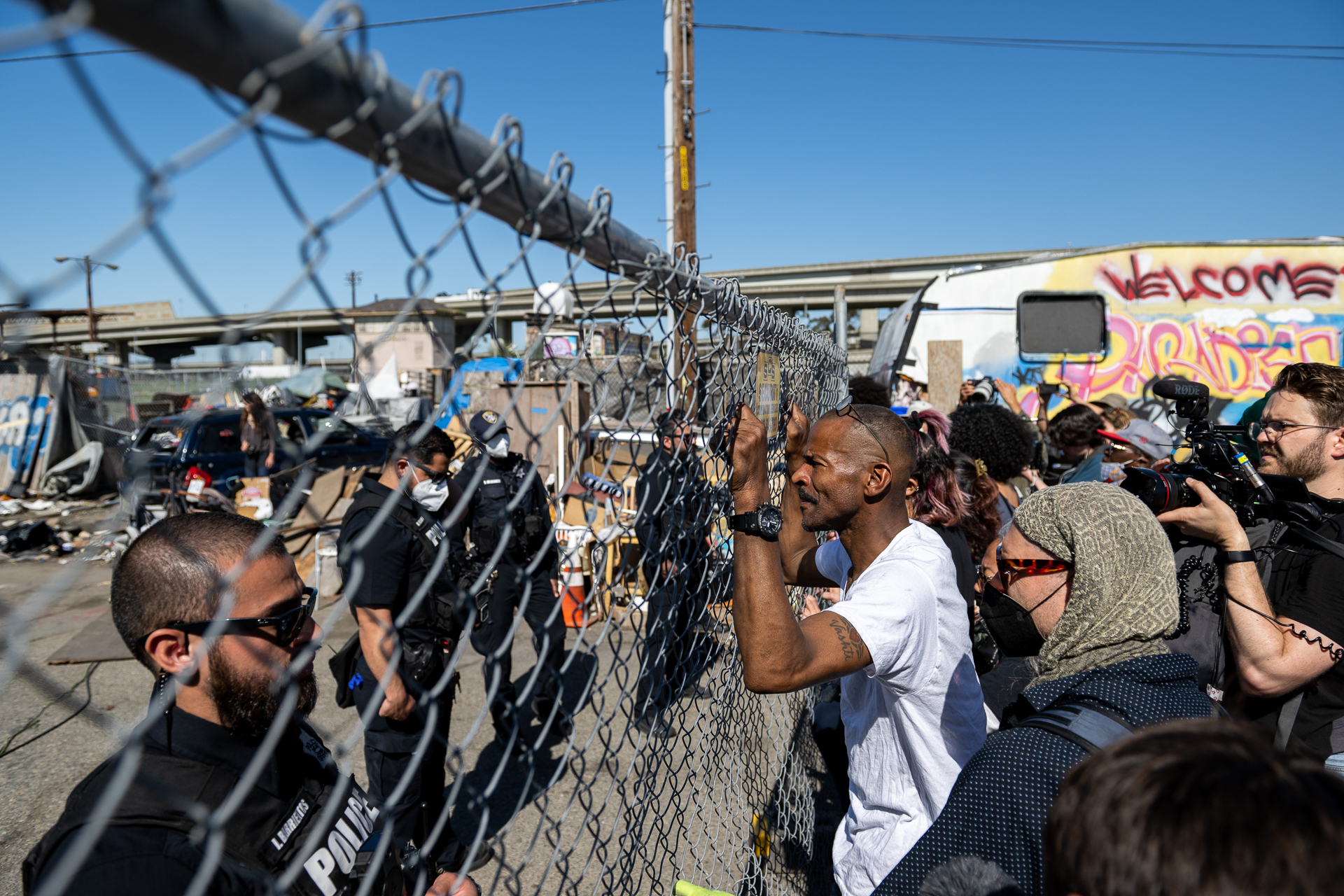 A man looks through a chain link fence at police officers on the other side of it. The man has a crowd of people behind him as his fingers rest in between the fence coils.